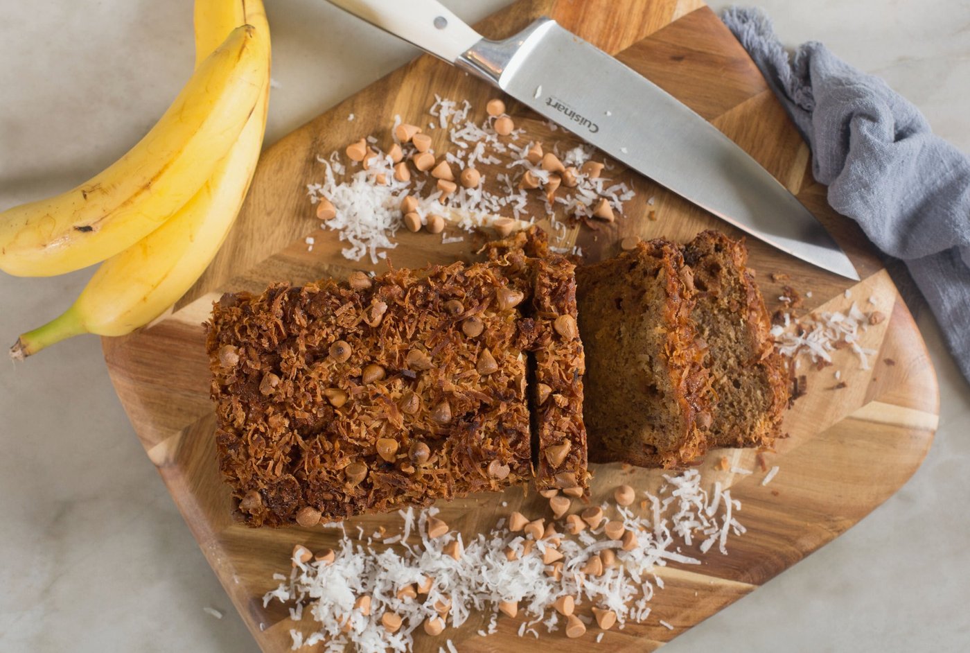 Severance Baking Co. coconut butterscotch banana bread on a wooden cutting board, surrounded by bananas, toasted coconut, and butterscotch chips